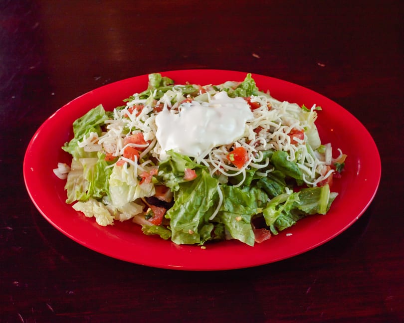 A vibrant red plate holds a fresh salad topped with shredded cheese, diced tomatoes, and a dollop of sour cream.