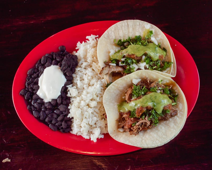Two tacos with meat, cilantro, onions, and green sauce, served with rice and black beans.