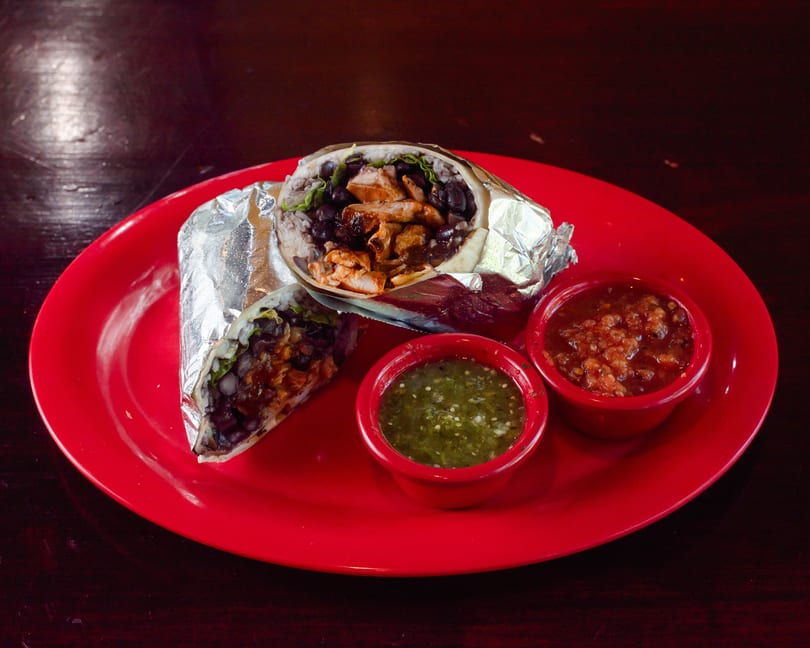 Two foil-wrapped burritos, one cut open, filled with chicken, black beans, and rice, served with two salsas on a red plate.