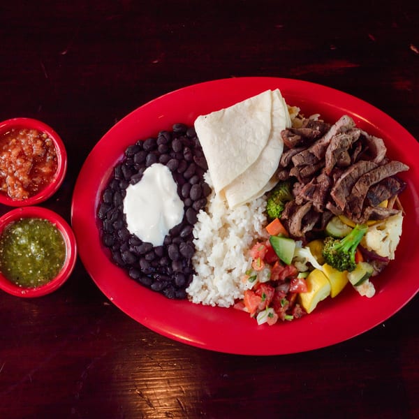 A vibrant red plate holds a meal with steak, rice, black beans, vegetables, tortillas, and two salsas.