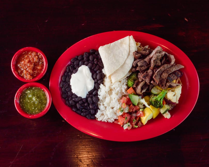 A vibrant red plate holds a meal with steak, rice, black beans, vegetables, tortillas, and two salsas.